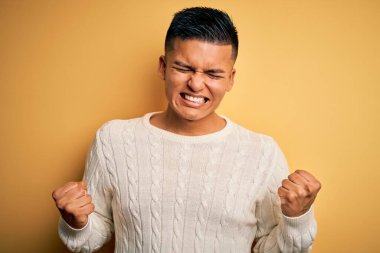 Sarı arka plan üzerine beyaz süveter giyen genç, yakışıklı, Latin bir adam çok mutlu ve heyecanlı bir şekilde kollarını kaldırıp, gülümseyerek ve başarı için bağırarak kazanan bir jest yapıyor. Kutlama konsepti.