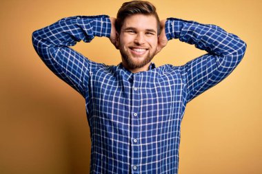 Sarışın, sakallı ve mavi gözlü genç bir işadamı sarı arka planda gömlek giyiyor rahatlıyor ve esniyor, kolları ve elleri başının arkasında ve boynunun arkasında mutlu bir şekilde gülümsüyor.