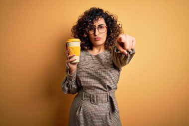 Genç, kıvırcık Arap iş kadını, gözlüklü, kahve bardağı içen, kamerayı işaret eden, el işareti, ön taraftan olumlu ve kendinden emin bir jest.