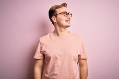 Genç, kızıl saçlı, günlük tişört giyen, izole edilmiş pembe arka plan üzerinde duran yüzünde gülümseme, doğal bir ifade ile yan yana bakan yakışıklı bir adam. Kendinden emin gülen.