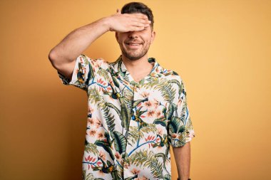 Tatilde mavi gözlü genç adam sarı arka planda çiçekli bir tişört giyiyor gülümsüyor ve elleri yüzünde şaşkınlıktan gözleri kapatıyordu. Kör kavram.