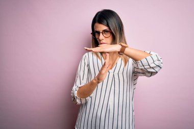 Genç ve güzel bir kadın, pembe arka planda günlük çizgili t-shirt ve gözlük takıyor. Elleriyle jest yapıyor, hayal kırıklığına uğramış ve ciddi bir yüz ifadesi var.