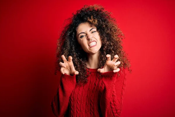 Young beautiful woman with curly hair and piercing wearing casual red sweater smiling funny doing claw gesture as cat, aggressive and sexy expression