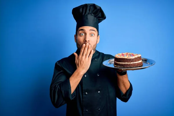 Young handsome cooker man with beard wearing uniform and hat holding tray with cake cover mouth with hand shocked with shame for mistake, expression of fear, scared in silence, secret concept