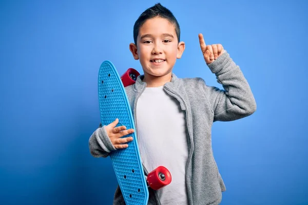 小男孩滑板手抱着现代滑板在蓝色孤立的背景下惊讶的一个想法或问题手指与快乐的脸 排名第一 — 图库照片