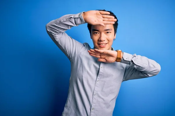 Young handsome chinese man wearing casual shirt standing over isolated blue background Smiling cheerful playing peek a boo with hands showing face. Surprised and exited