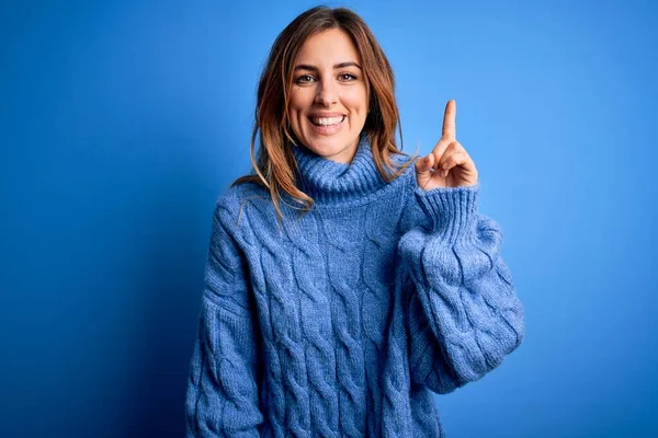 Young beautiful brunette woman wearing casual turtleneck sweater over blue background showing and pointing up with finger number one while smiling confident and happy.