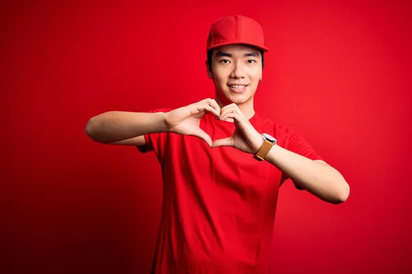 Young handsome chinese delivery man wearing cap standing over isolated red background smiling in love showing heart symbol and shape with hands. Romantic concept.