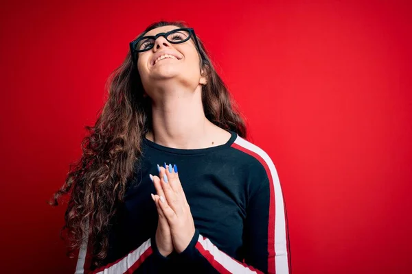 Young beautiful woman with curly hair wearing sweater and glasses over red background begging and praying with hands together with hope expression on face very emotional and worried. Begging.