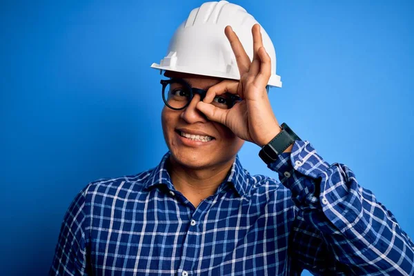 Young handsome engineer latin man wearing safety helmet over isolated blue background doing ok gesture with hand smiling, eye looking through fingers with happy face.