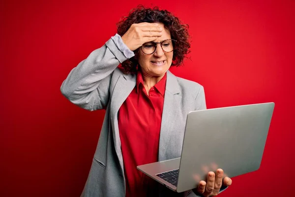 中年卷发女商人用笔记本电脑在孤立的红色背景下工作时 手牵着手 满脸羞愧和惊讶 愤怒和沮丧 恐惧和因错误而烦恼 — 图库照片