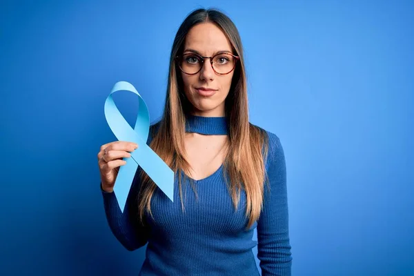 Young blonde woman with blue eyes holding colon cancer awareness blue ribbon with a confident expression on smart face thinking serious