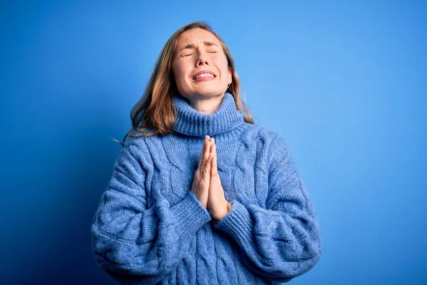Young beautiful blonde woman wearing casual turtleneck sweater over blue background begging and praying with hands together with hope expression on face very emotional and worried. Begging.