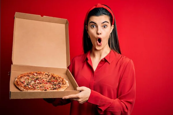 若いです美しいですブルネット女性保持デリバリーボックスとともにイタリアのピザ以上赤背景怖がってショックで驚きの顔 恐怖と興奮で表現 — ストック写真
