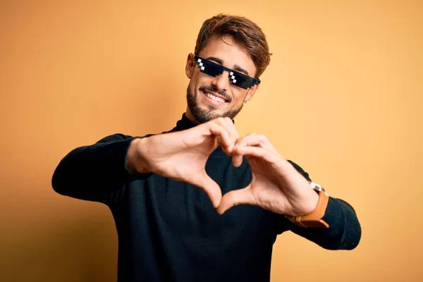Young man wearing thug life fanny sunglasses standing over isolated yellow background smiling in love showing heart symbol and shape with hands. Romantic concept.