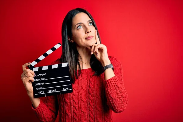 Young director woman with blue eyes making movie holding clapboard over red background serious face thinking about question, very confused idea