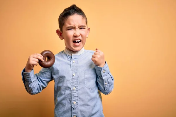 Młody Chłopiec Jedzący Niezdrowe Pączki Czekoladowe Odizolowanym Żółtym Tle Zirytowany — Zdjęcie stockowe