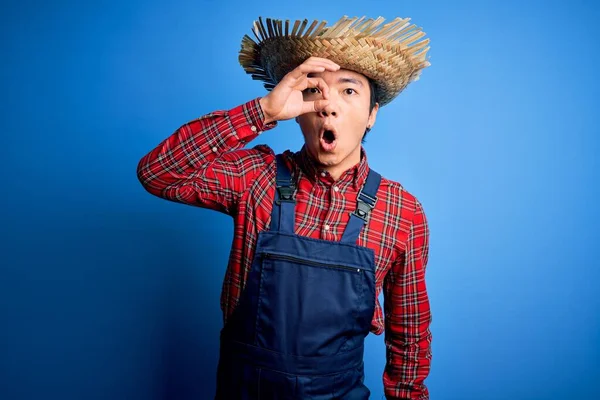 Young handsome chinese farmer man wearing apron and straw hat over blue background doing ok gesture shocked with surprised face, eye looking through fingers. Unbelieving expression.