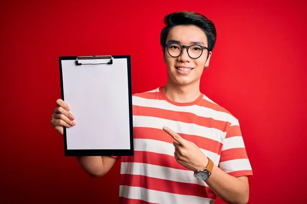 Young handsome chinese inspector man wearing glasses holding clipboard checklist very happy pointing with hand and finger