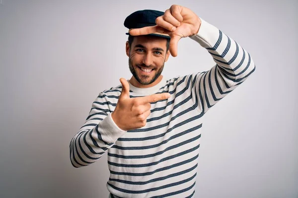 Young handsome sailor man with beard wearing navy striped uniform and captain hat smiling making frame with hands and fingers with happy face. Creativity and photography concept.