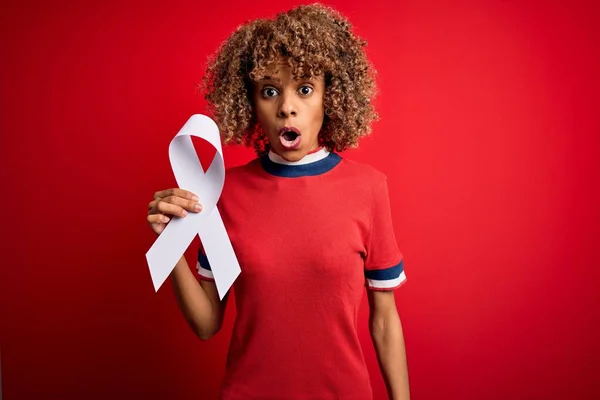 Young african american woman with curly hair holding white cancer ribbon over red background scared in shock with a surprise face, afraid and excited with fear expression