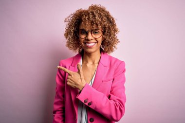 Genç Afro-Amerikalı iş kadını pembe arka planda duran gözlüklü neşeli bir gülümsemeyle mutlu ve doğal bir ifadeyle elini ve parmağını kaldırdı.