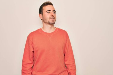 Mavi gözlü genç bir adam beyaz arka planda dikilip yüzünde gülümseme, doğal bir ifade ile yan yana duran sıradan bir kazak giyiyor. Kendinden emin gülen.