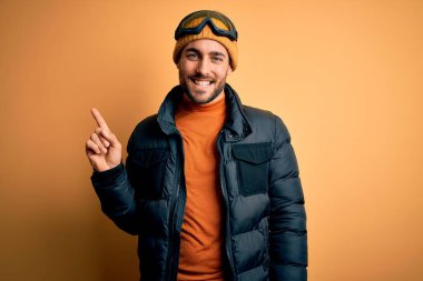 Sakallı, kar maskesi takan ve yüzünde kocaman bir gülümsemeyle kayak gözlüğü takan yakışıklı bir kayakçı. Kameraya el parmağı ile bakıyor..