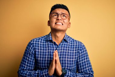 Sarı arka plan üzerine gündelik tişört ve gözlük takan genç, yakışıklı Latin bir adam elleri bağlı yalvarıyor ve dua ediyor. Yüzünde umut ifadesi var. Çok duygusal ve endişeli. Yalvarıyorum..