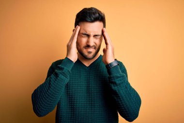 Sakallı genç yakışıklı adam sarı arka planda dikiliyor. Ağrı ve migren yüzünden baş ağrısı çekiyor. Ellerini başının üstüne koy..