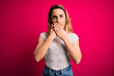 İzole edilmiş pembe arka planda duran gündelik tişört giyen genç ve güzel sarışın kadın yanlışlıkla ağızlarını kapatarak şok geçirdi. Gizli konsept.