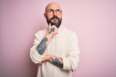 Sakallı, dövmeli, gözlüklü, pembe arka planda süveteri olan, eli çenesinde soru soran yakışıklı kel bir adam. Düşünceli bir ifade. Düşünceli bir yüzle gülümsüyor. Şüphe kavramı.