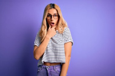 Mor arka planda çizgili t-shirt ve gözlük takan genç, güzel sarışın kadın elleri çenesinde olan inançsızlıktan, şaşkınlıktan ve şaşkınlıktan büyülenmiş görünüyor.