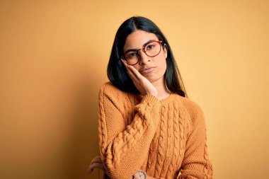 Sarı arka plan üzerine gözlük takan genç ve güzel İspanyol kadın çapraz kollu depresyondan sıkılmış ve yorgun görünüyor..