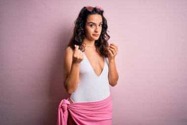 Tatilde kıvırcık saçlı güzel bir kadın pembe arka planda beyaz mayo giyer, para jesti yapar, maaş ödemesi ister, milyonerlik işi yapar.