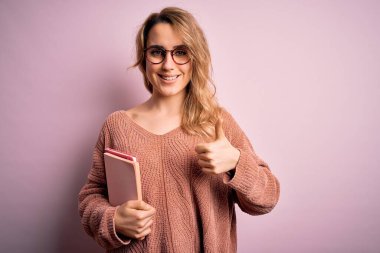 Pembe arka planda gözlük takan güzel sarışın kadın mutlu bir şekilde gülümsüyor tamam işareti yapıyor, parmaklarıyla başparmağını kaldırıyor, mükemmel bir işaret.