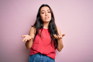 Genç esmer kadın pembe izole edilmiş arka plan üzerine gündelik bir tişört giyiyor ve kolları açık, hiçbir fikri yok..