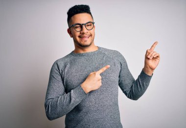 İzole edilmiş beyaz arka planda gözlük ve süveter giyen genç yakışıklı adam gülümsüyor ve kameraya bakıyor iki eliyle ve parmaklarıyla kenarı işaret ediyor..