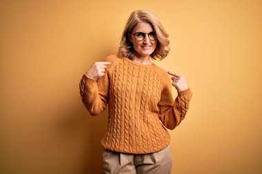 Orta yaşlı güzel sarışın kadın sarı arka planda günlük süveter ve gözlük takıyor. Kendine güler yüzlü, gururlu ve mutlu birini işaret ediyor..