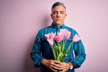 Middle age grey-haired man holding natural bouquet of pink tulips over isolated background with a confident expression on smart face thinking serious clipart