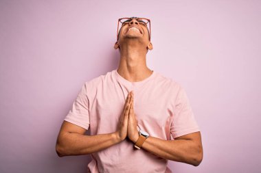 Yakışıklı Afro-Amerikan erkeği pembe arka planda günlük tişört ve gözlük takıyor, yalvarıyor ve dua ediyor. Yüzünde umut ifadesi olan çok duygusal ve endişeli bir ifade var. Yalvarıyorum..