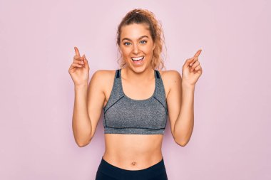 Genç, sarışın, mavi gözlü sporcu kadın spor kıyafeti giyiyor. Gülümsüyor, şaşırıyor, parmaklarıyla işaret ediyor ve kollarını kaldırıyor..