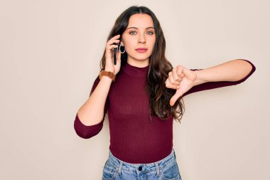 Genç ve güzel bir kadın beyaz arka planda akıllı telefondan konuşuyor. Kızgın bir yüzü var. Negatif işaretler baş parmakları aşağıda, reddedilme konsepti ise reddedilmiş.