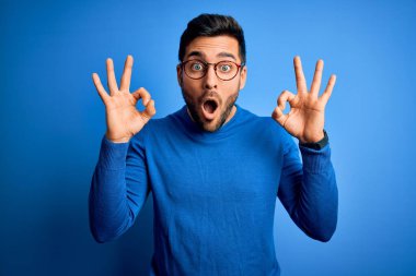 Mavi arka planda günlük süveter ve gözlük takan sakallı yakışıklı genç adam şaşırmış ve şaşırmış görünüyor. Parmaklarıyla onay sembolü yapıyor. Çılgın ifade