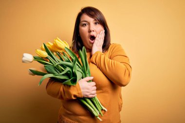 Sarı arka planda romantik lale çiçekleri tutan güzel ve iri bir kadın. Korkmuş ve şok olmuş, şaşırmış ve şaşırmış bir yüz ifadesi var.