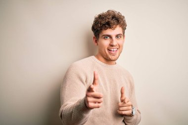 Young blond handsome man with curly hair wearing casual sweater over white background pointing fingers to camera with happy and funny face. Good energy and vibes.