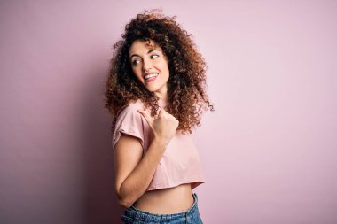 Kıvırcık saçlı ve piercing giyen mutlu yüzüyle gülümseyen ve baş parmağıyla yan tarafı gösteren sıradan pembe tişörtlü güzel bir kadın..