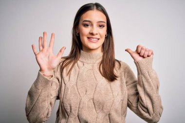 Genç, güzel, esmer, sıradan bir süveter giyen, beyaz arka plan üzerinde duran, altı numarayı gösteren, kendinden emin ve mutlu bir şekilde gülümseyen bir kadın..