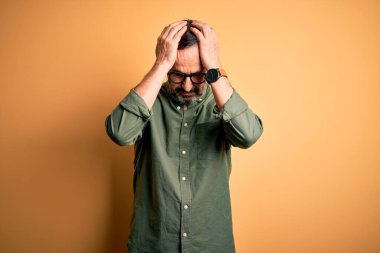 Orta yaşlı, beyaz, günlük yeşil gömlek ve gözlük takan, izole edilmiş sarı arka planda baş ağrısı çeken, çaresiz ve stresli bir adam. Çünkü ağrı ve migren. Ellerini başının üstüne koy..