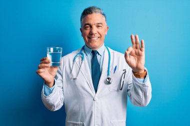 Orta yaşlı, yakışıklı, gri saçlı doktor. Mavi arka planda bir bardak su içiyor. Parmaklarıyla imza atıyor.